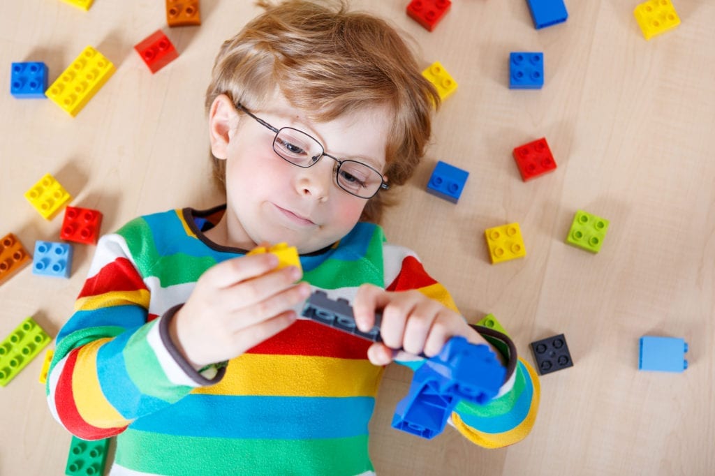 Little blond kid boy playing with lots of colorful plastic blocks indoor. child wearing colorful shirt and glasses, having fun with building and creating.