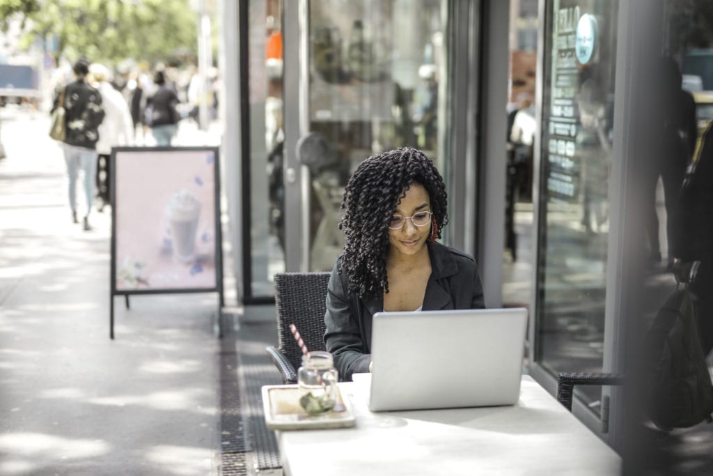 Ethnic young woman using laptop while having tasty beverage in modern street cafe