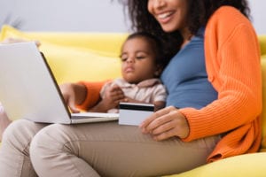 happy african american mother holding credit card and using laptop near toddler daughter in living room