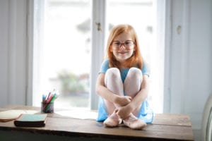 Happy cute small girl in glasses sitting on table in light living room