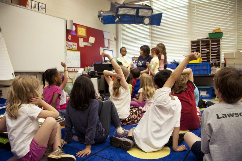 Captured in a metropolitan Atlanta, Georgia primary school, this photograph depicts a typical classroom scene, where an audience of school children were seated on the floor before a teacher at the front of the room, who was reading an illustrated storybook, during one of the scheduled classroom sessions. Assisting the instructor were two female students to her left, and a male student on her right, who was holding up the book, while the seated classmates were raising their hands to answer questions related to the story just read.