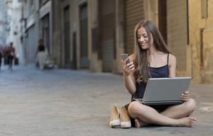 A woman sitting on the floor and using her phone and laptop.