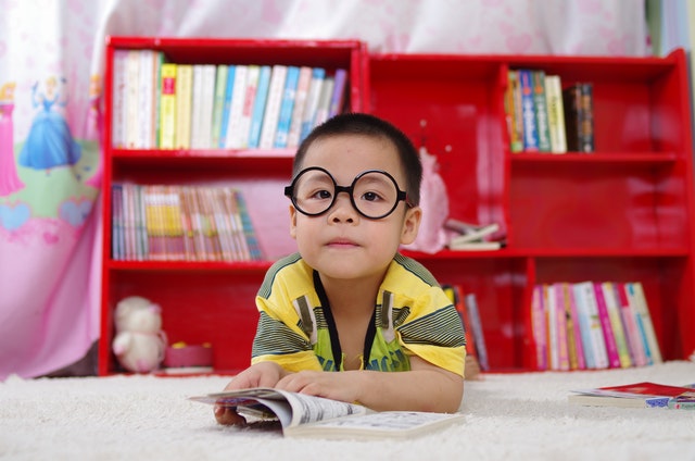 A red shelf with a lot of books behind a boy with glasses sitting at a desk and reading a book