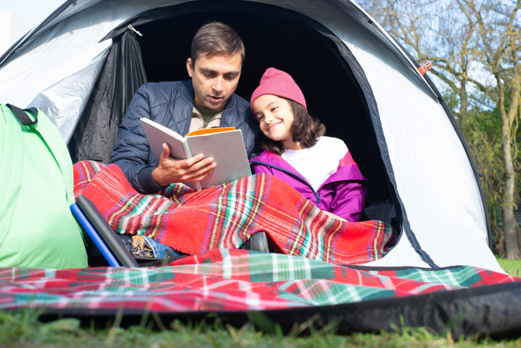 Father and daughter reading a book while sitting inside the tent